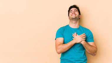 Bej arka planda izole olmuş genç adam gülüyor, ellerini kalbe koyuyor, mutluluk kavramını koruyor..