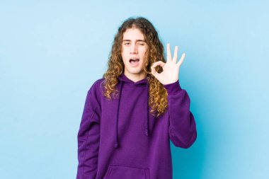 Uzun saçlı adam yalnız poz veriyor bir göz kırpıyor ve elleriyle iyi bir jest yapıyor..