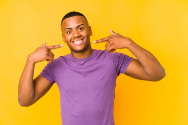 Sarı arka planda izole edilmiş genç latin adam gülümsüyor, parmaklarını ağıza doğrultuyor..