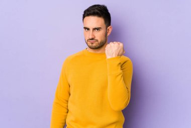 Mor arka planda izole edilmiş genç beyaz adam yumruğunu kameraya gösteriyor, agresif yüz ifadesi var..