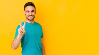 Sarı fırında izole edilmiş genç beyaz adam zafer işareti gösteriyor ve gülümsüyor..