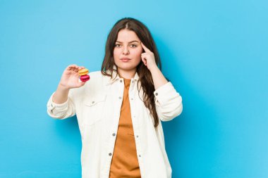 Elinde badem kurabiyesi tutan, parmağıyla şakağı işaret eden, düşünen, bir göreve odaklanmış, kıvrımlı bir kadın..