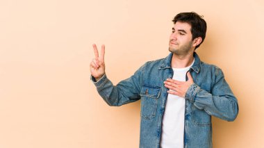 Genç adam bej arka planda izole edilmiş. Yemin ediyor, elini göğsüne koyuyor..