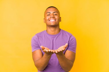 Sarı arka planda izole edilmiş genç latin adam elinde avuç içi bir şeyle kameraya sunuyor..