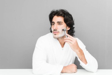 Gri bir arka planda sakalını kesen genç beyaz bir adam.