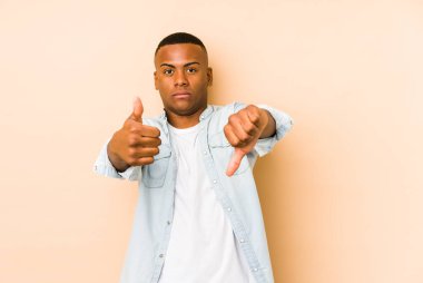 Genç latin adam bej arka planda izole edilmiş. Baş parmaklarını yukarı ve baş parmaklarını aşağı gösteriyor. Zor seçim konsepti.