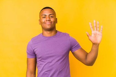 Sarı arka planda izole edilmiş genç latin adam gülümseyerek beş numarayı parmaklarıyla gösteriyor..