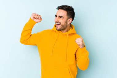 Mavi arka planda izole genç beyaz adam özel bir günü kutluyor, atlıyor ve enerjiyle kollarını kaldırıyor..