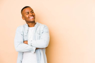 Bej arka planda izole edilmiş genç Latin adam kollarını kavuşturarak gülümsüyor..