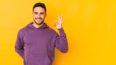 Kafkasyalı genç adam sarı bir alanda izole edilmiş bir şekilde bir göz kırpıyor ve elleriyle iyi bir jest yapıyor..