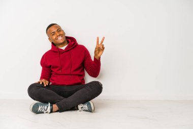 Yerde oturan genç latin adam neşeli ve tasasız bir şekilde parmaklarıyla barış sembolü gösteriyor..