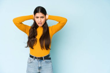 Genç Hintli bir kadın mavi arka planda izole edilmiş kulaklarını elleriyle kapatarak yüksek sesli sesler duymamaya çalışıyor..