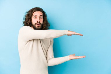 Uzun saçlı genç adam mavi arka planda izole edilmiş. Elleri arasında bir kopya alanı tutarken şok ve hayret içinde..