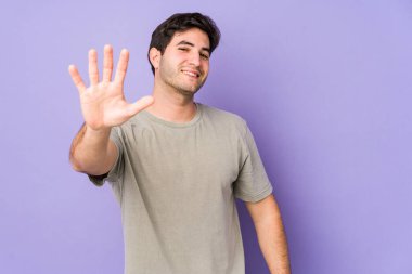 Mor arka planda izole edilmiş genç adam gülümseyerek beş numarayı parmaklarıyla gösteriyor..