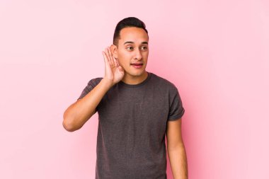 Pembe arka planda poz veren Latin bir adam bir dedikoduyu dinlemeye çalışıyor..