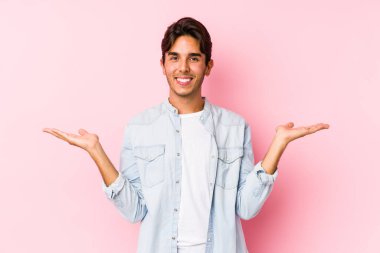 Pembe arka planda poz veren genç beyaz adam kollarıyla ölçek oluşturuyor, mutlu ve kendinden emin hissediyor..