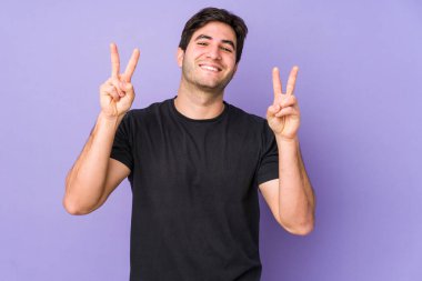 Young man isolated on purple background showing victory sign and smiling broadly. clipart