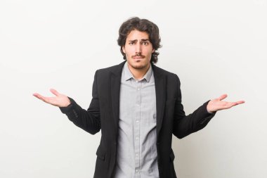 Genç bir iş adamı beyaz bir arkaplanda, kafası karışmış ve omuz silkiyor..