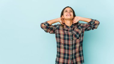 Mavi arka planda izole edilmiş genç bir kadın kendini güvende hissediyor, elleri başının arkasında..