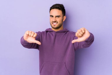 Mor arka planda izole edilmiş genç beyaz adam baş parmağını aşağı gösteriyor ve hoşlanmadığını ifade ediyor..