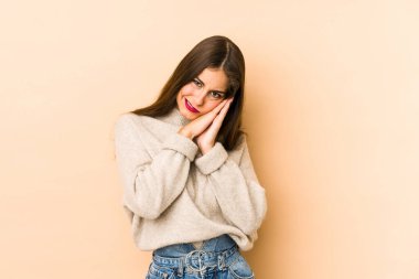 Beyazlı genç bir kadın bej arka planda izole edilmiş esniyor. Ağzı eliyle kapatan yorgun bir hareket sergiliyor..