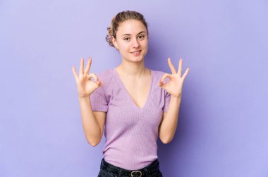 Mor arka planda genç, beyaz bir kadın neşeli ve kendinden emin, iyi bir jest yapıyor..