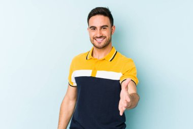 Mavi arka planda izole edilmiş genç beyaz adam selamlama jesti olarak kameraya elini uzatıyor..