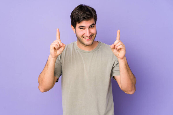 Young man isolated on purple background indicates with both fore fingers up showing a blank space.