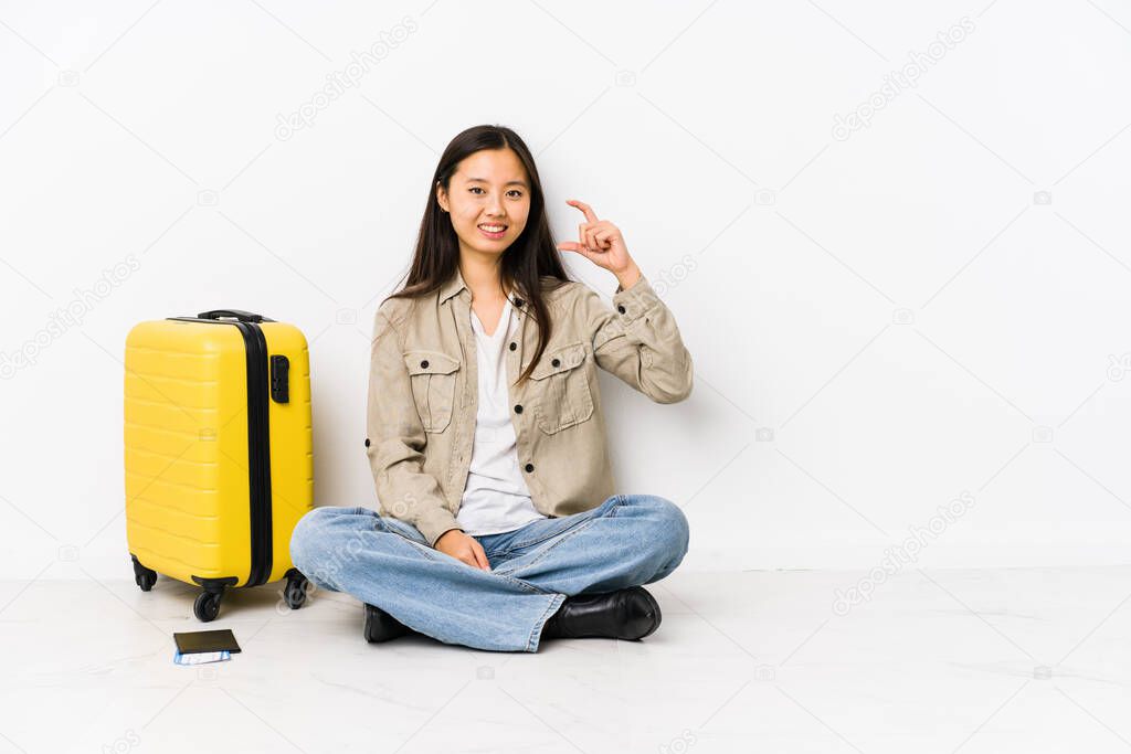 Young chinese traveler woman sitting holding a boarding passes holding something little with forefingers, smiling and confident.