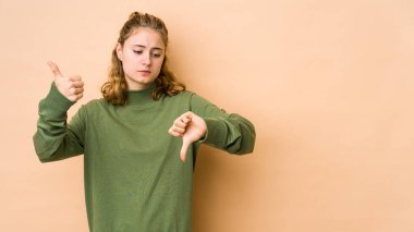 Beyaz tenli genç bir kadın bej arka planda izole edilmiş. Baş parmağını kaldırmış, baş parmağını aşağı gösteriyor. Zor seçim konsepti.