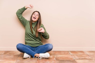 Genç beyaz kadın yerde tek başına oturmuş özel bir günü kutluyor, atlıyor ve enerjiyle kollarını kaldırıyor..