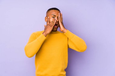 Mor arka planda izole edilmiş genç latin adam parmaklarının arasından korkmuş ve gergin bir şekilde göz kırp..