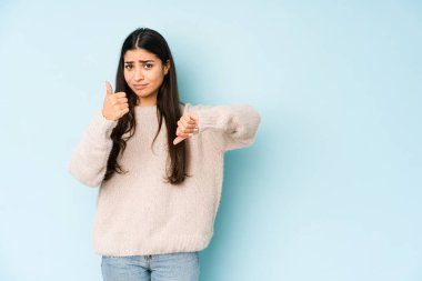 Mavi arka planda izole edilmiş genç Hintli kadın baş parmağını kaldırıyor, zor seçim konsepti.