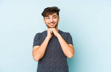 Mavi arka planda izole edilmiş genç bir Arap adam ellerini çenesinin altında tutuyor, mutlu bir şekilde yana bakıyor..