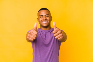 Sarı arka planda izole olmuş genç latin bir adam. Başparmakları havada, bir şey için tezahürat yapıyor. Destek ve saygı kavramı..