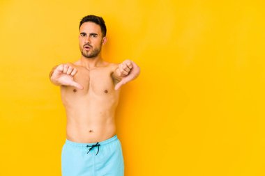 Sarı arka planda mayosu olan genç, beyaz bir adam. Başparmağını öne eğip, hoşlanmadığını ifade eden beyaz bir adam..