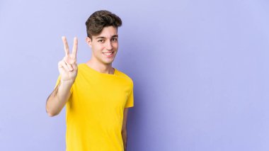 Mor arka planda izole edilmiş genç beyaz adam zafer işareti gösteriyor ve gülümsüyor..