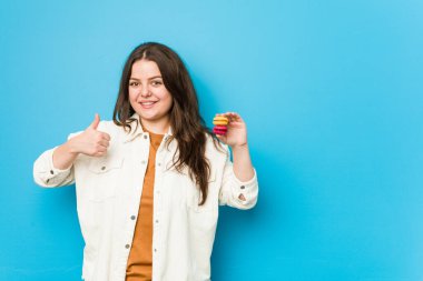 Kurabiye tutan, gülümseyen ve baş parmağını kaldıran kıvrımlı genç bir kadın.