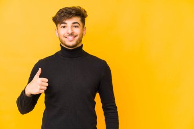 Sarı arka planda izole edilmiş genç bir Arap adam gülümsüyor ve başparmağını kaldırıyor.