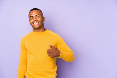 Mor arka planda izole olmuş genç latin adam gülümsüyor ve baş parmağını kaldırıyor.