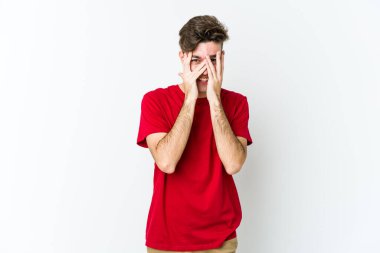 Beyaz arka planda izole edilmiş genç beyaz bir adam kameraya parmaklarıyla göz kırpıyor, yüzünü kapatmaktan utanıyor..