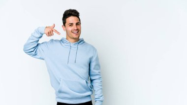 Beyaz arka planda yaşayan beyaz tenli genç bir adam gömlek fotokopi alanını gösteriyor, gururlu ve kendinden emin.