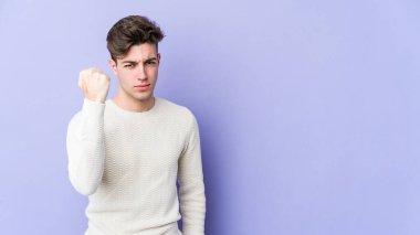 Mor arka planda izole edilmiş genç beyaz adam yumruğunu kameraya gösteriyor, agresif yüz ifadesi var..