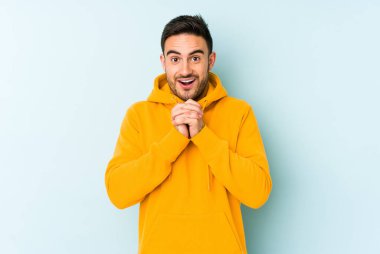 Kafkasyalı genç adam mavi arka planda izole edilmiş şans için dua ediyor, şaşırmış ve ağzını açıp önüne bakıyor..