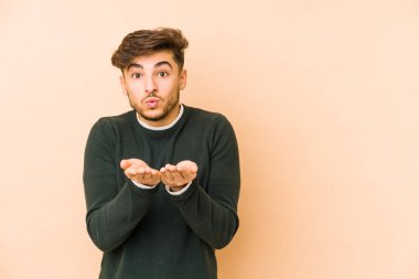 Genç Arap bir adam bej bir arka planda izole edilmiş dudaklar kıvrılmış ve avuç içleriyle hava öpücüğü yolluyor..