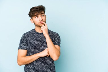 Mavi arka planda izole edilmiş genç bir Arap şüpheli ve şüpheci bir ifadeyle yana bakıyor..