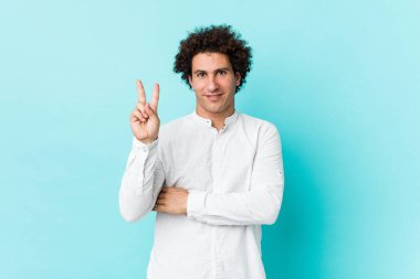 Genç, kıvırcık, olgun bir adam zarif bir gömlek giyiyor. İki numarayı parmaklarıyla gösteriyor..