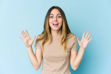 Mavi arka planda izole genç beyaz kadın hoş bir sürpriz alıyor, heyecanlı ve el kaldırıyor..