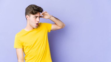 Mor arka planda izole edilmiş genç beyaz adam uzaklara bakıyor ve elini alnına koyuyor..