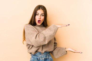 Beyazlı genç bir kadın bej arka planda izole edilmiş. Elleri arasında bir kopya alanı tutarken şok olmuş ve hayrete düşmüştü..